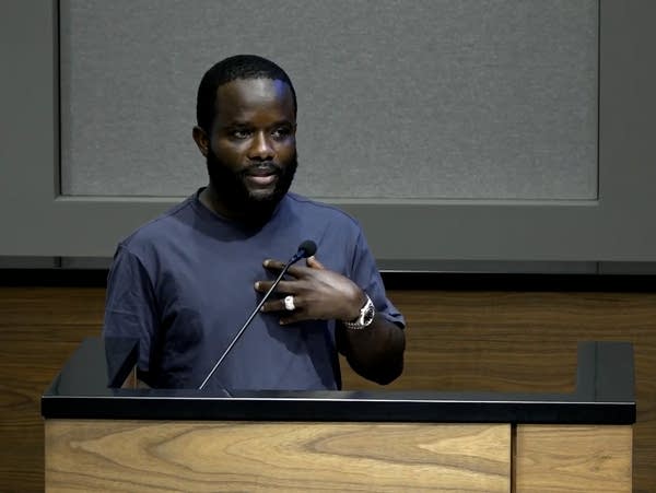A man touches his hand to his chest while speaking at a podium.