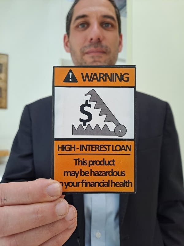 Özgür Ince, a professor of finance, wears a black suit. He is holding a small orange sign that reads "Warning! HIGH-INTEREST LOAN. This product may be hazardous to your financial health." In a small white box, a bear trap closes in around a dollar sign.