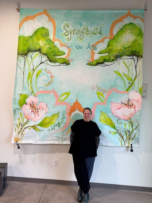 A woman stands in front of a sign that reads "Springboard For The Arts" and natural paintings.