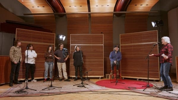 Seven people have a conversation in a recording studio