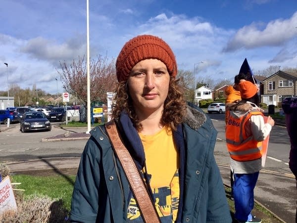 Dr. Naomi Dunne, who has shoulder-length curly hair and wears an orange shirt, blue jacket and orange beanie, stands in a parking lot and faces the camera with a serious expression on her face.