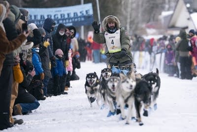 A dog sled races past a crowd of spectators while the musher waves.