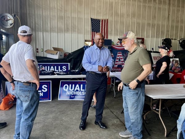 A political candidate speaks with attendees in front of campaign signs at a picnic.