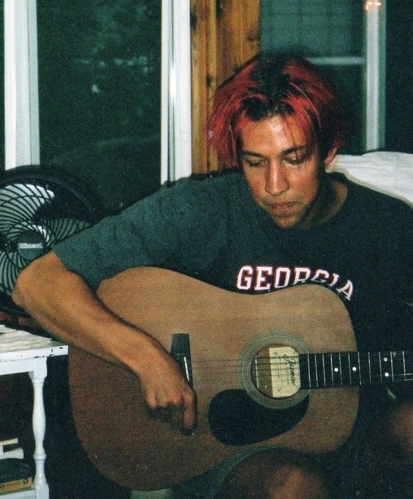 A man with his hair dyed pink strums an acoustic guitar