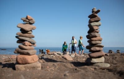 People walking along a lakeshore are framed by stacks of stones. 