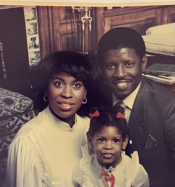 An older-looking family photo with a Black woman in a white shirt, a Black man in a dark suit, and a young Black girl in a white shirt.