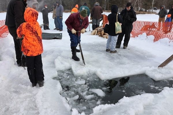 A man uses a long hand saw to carve ice while surrounded by people.