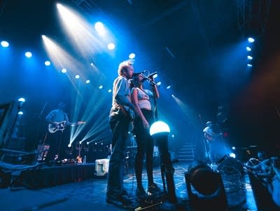 A woman sings into a microphone next to a man with a bass singing into a microphone on a lighted stage.