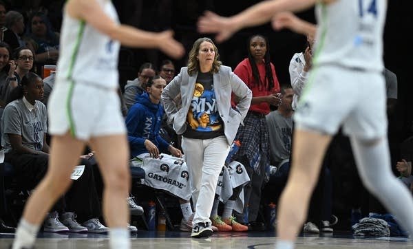 A basketball coach is seen on the sidelines with her hands in her pockets as players are seen in the foreground.