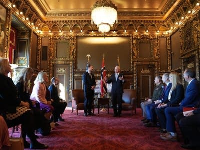 Two men speak at the front of a room while other people are gathered along the sides