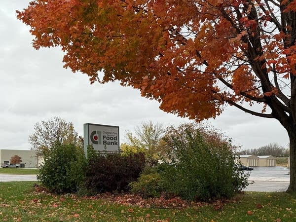A sign for a food bank sits under a tree with orange leaves.