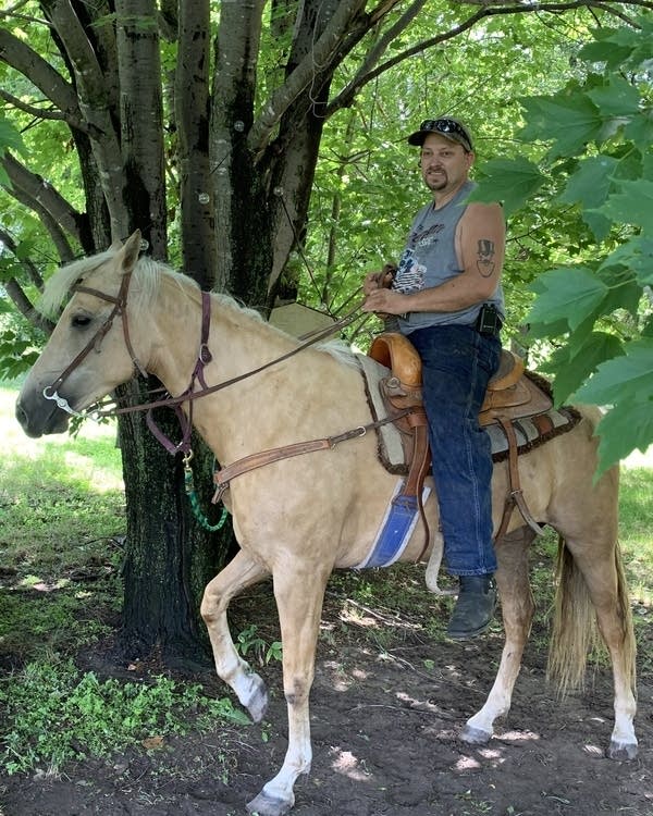 A man in a sleeveless gray T-shirt rides a light-tan horse.