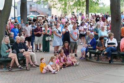 Wide shot of vigil attendees standing and sitting as they listen to a speaker.