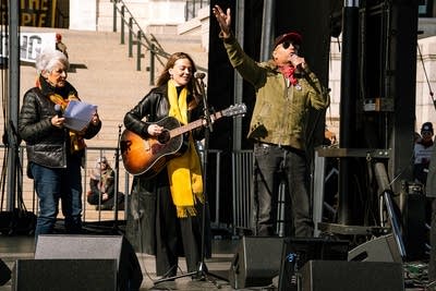 Three musicians perform together on an outdoor stage at a large rally