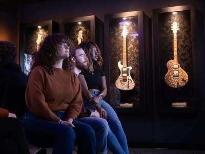 A group of people sit in wonder with a wall of electric guitars behind them.
