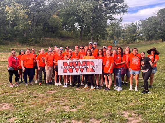 People in orange shirts pose for a group photo with a Native Women Run banner.