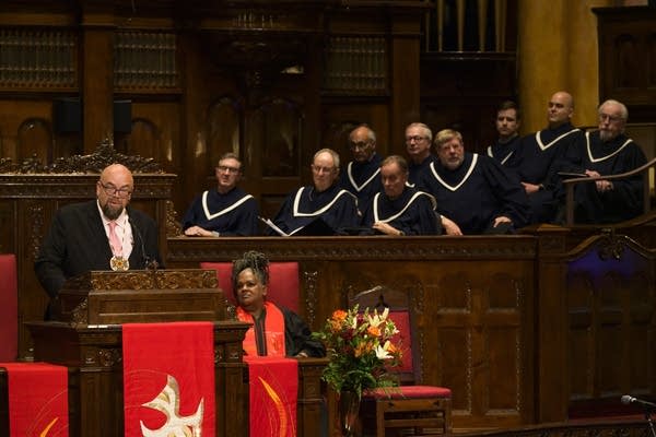 A man preaches at a pulpit with a choir behind him.