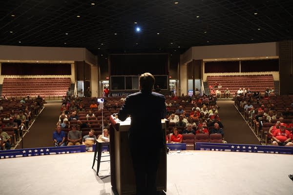 A shot of a politician from behind speaking on stage to an auditorium of people.