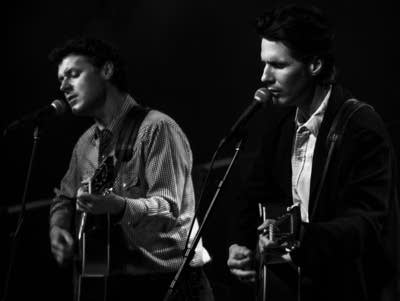 Two men sing into microphones and play acoustic guitars.