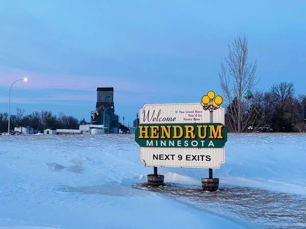 A sign reading "Welcome to Hendrum, Minnesota - Next 9 Exits" stands in the snow.