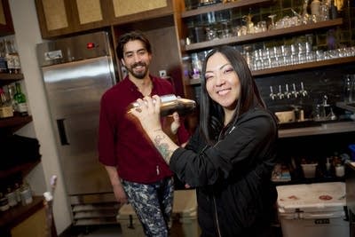 Two hospitality workers smile for a photo; one mixes a drink