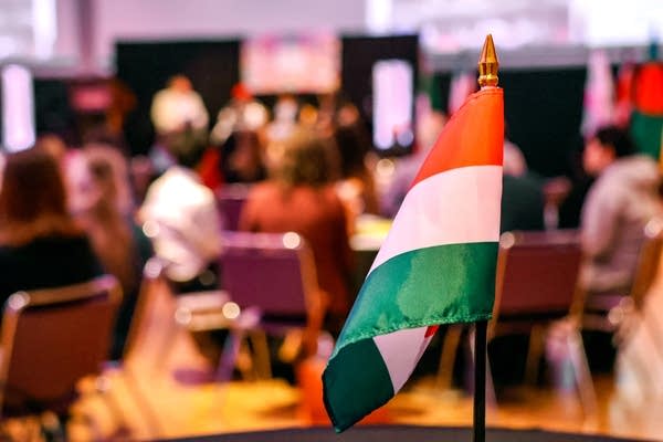 An orange, white and green flag is seen on a table in the foreground. In the background, people sit around tables in a banquet hall.