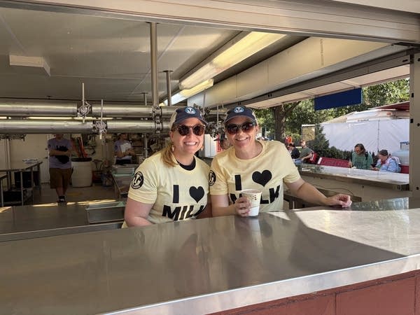 Two women lean together behind a steel counter to pose for a photo.