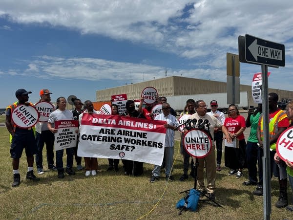 A man speaks at a podium outdoors next to a group of workers holding a sign that reads, 