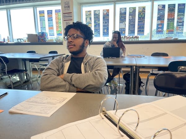 A student in a grey sweatshirt sits in a classroom with bright light coming in the window behind him. 