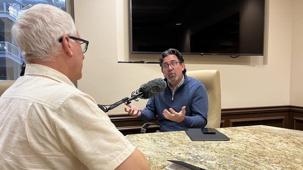 Jeremy Aguero of Applied Analysis sits at a marble table with a microphone in front of his face, speaking with "Marketplace Morning Report" host David Brancaccio.