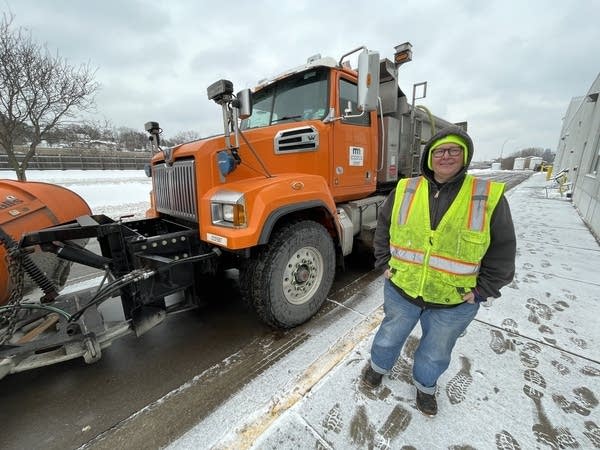 A woman in a hi-vis vest stands next to an orange snow plow on a street.