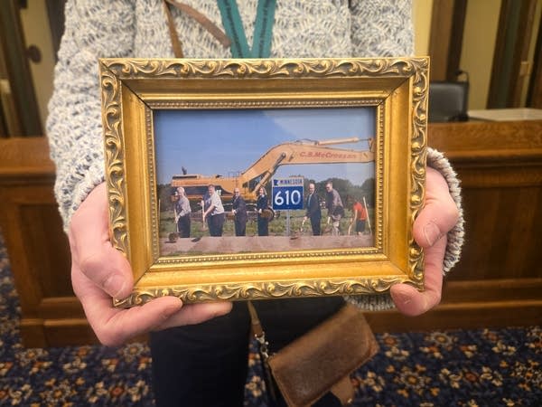 A woman in an office holds a golden framed photo of people breaking ground on a highway.