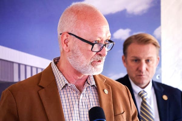 A man in glasses and a brown jacket speaks into a microphone.