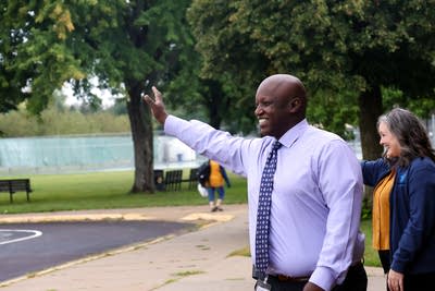 A man in a shirt and tie waves to students in a parking lot.