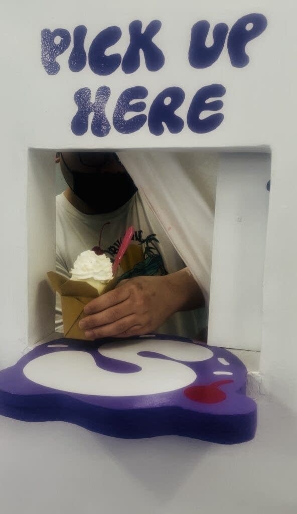 A hand pokes through a small square holding a cup of ice cream. A sign above it reads the hole in the wall reads "Pick Up Here."