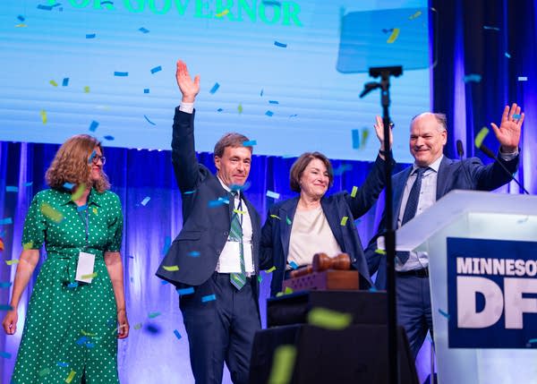 Four people on a stage three of are waving to a crowd