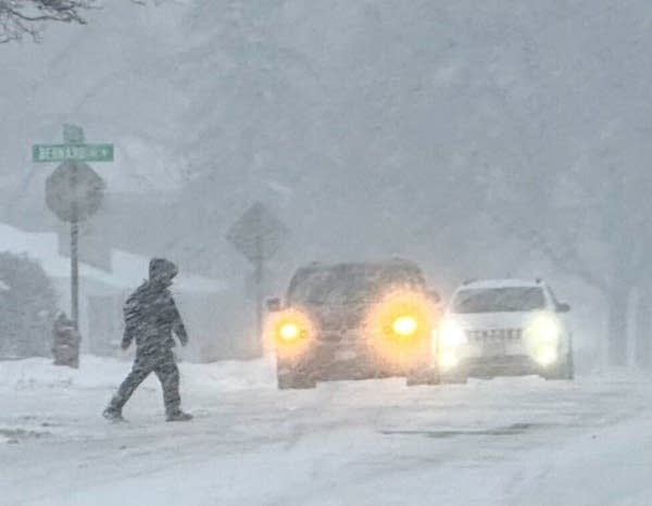 A person crosses a street in front of two cars during a snow storm.