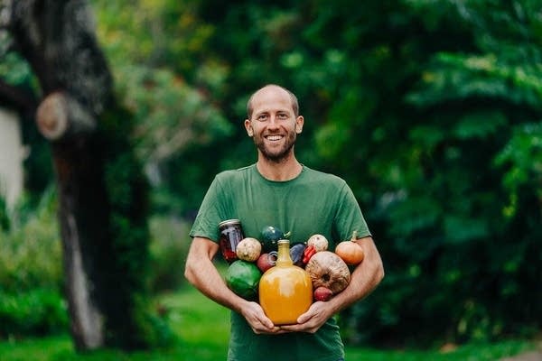 A man standing in a green forested area holds jars and vegetables.