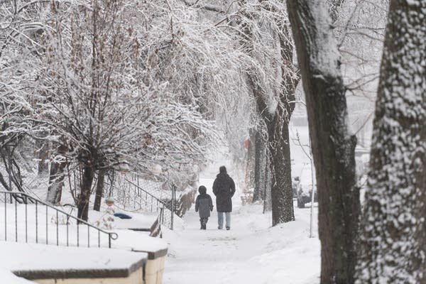 An adult and a child walk down a sidewalk during a snowstorm.