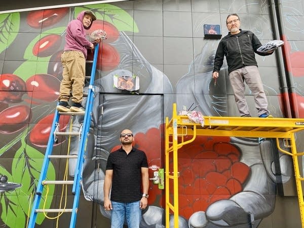 one man stands on a ladder, another stands on a scaffolding, and one stands on the ground.
