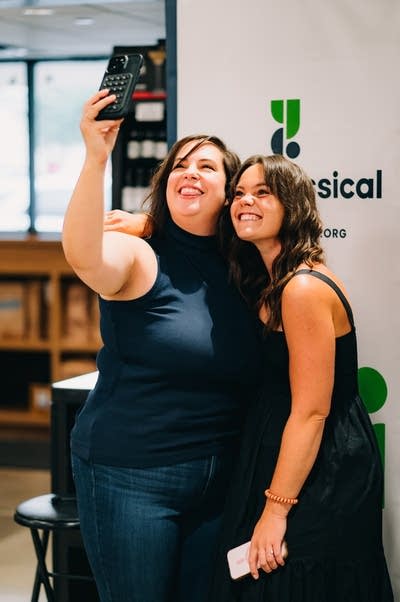 Two women stand together for a selfie at a social gathering inside a wine shop