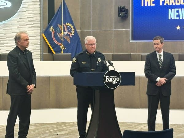 A police chief speaks at a podium while flanked by men in suits.