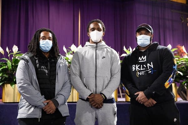 Three men stand in front of purple curtains.