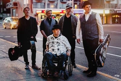 Five men gathered alongside an urban street, two with guitar cases 