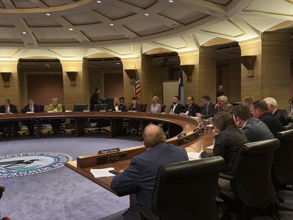 A group of people sit around a large wooden desk in a circular room.