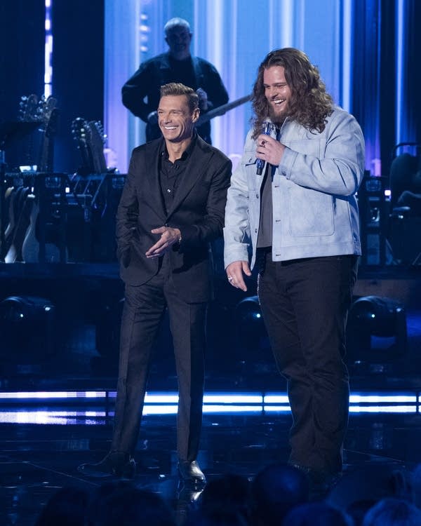 A man with long, curly hair sings smiles on stage next to a host in a black suit.