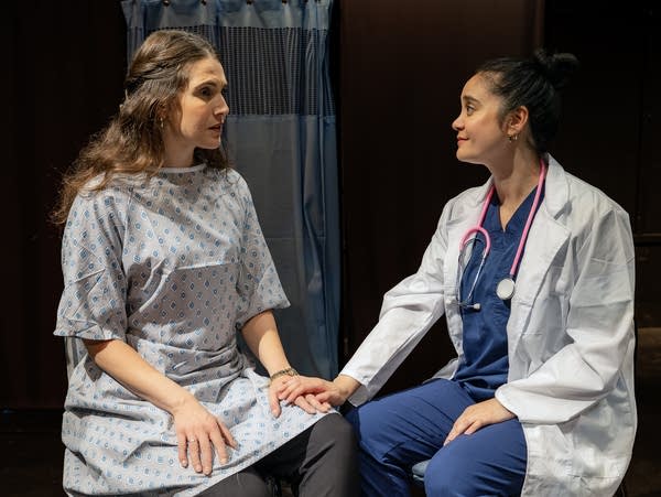 Actors portraying a patient and a doctor sit on a stage.