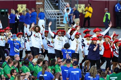Athletes in white shirts and red hats wave to people in a stadium.
