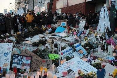 A group of people gather around a memorial full of signs, flowers and candles.