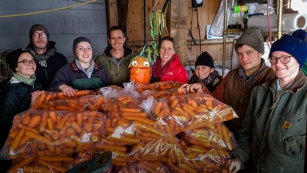 a group of people stand around stacks of bagged carrots